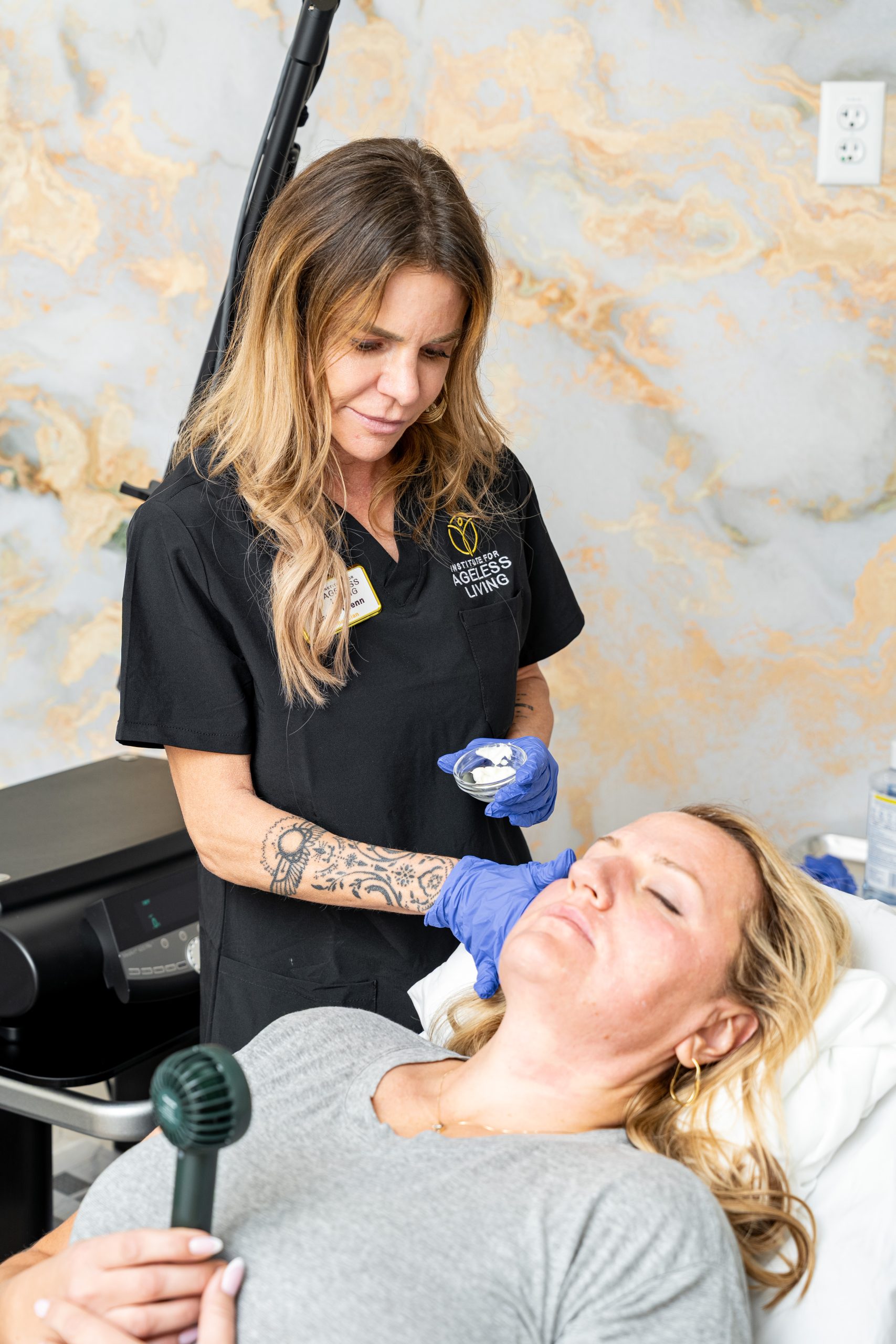 A woman wearing gloves and a black uniform prepares to treat a patient lying on a bed with her eyes closed, holding a jade roller. The serene setting, reminiscent of the Institute For Ageless Living, emphasizes both wellness and relaxation in what appears to be an advanced clinic or spa focused on anti-aging treatments.