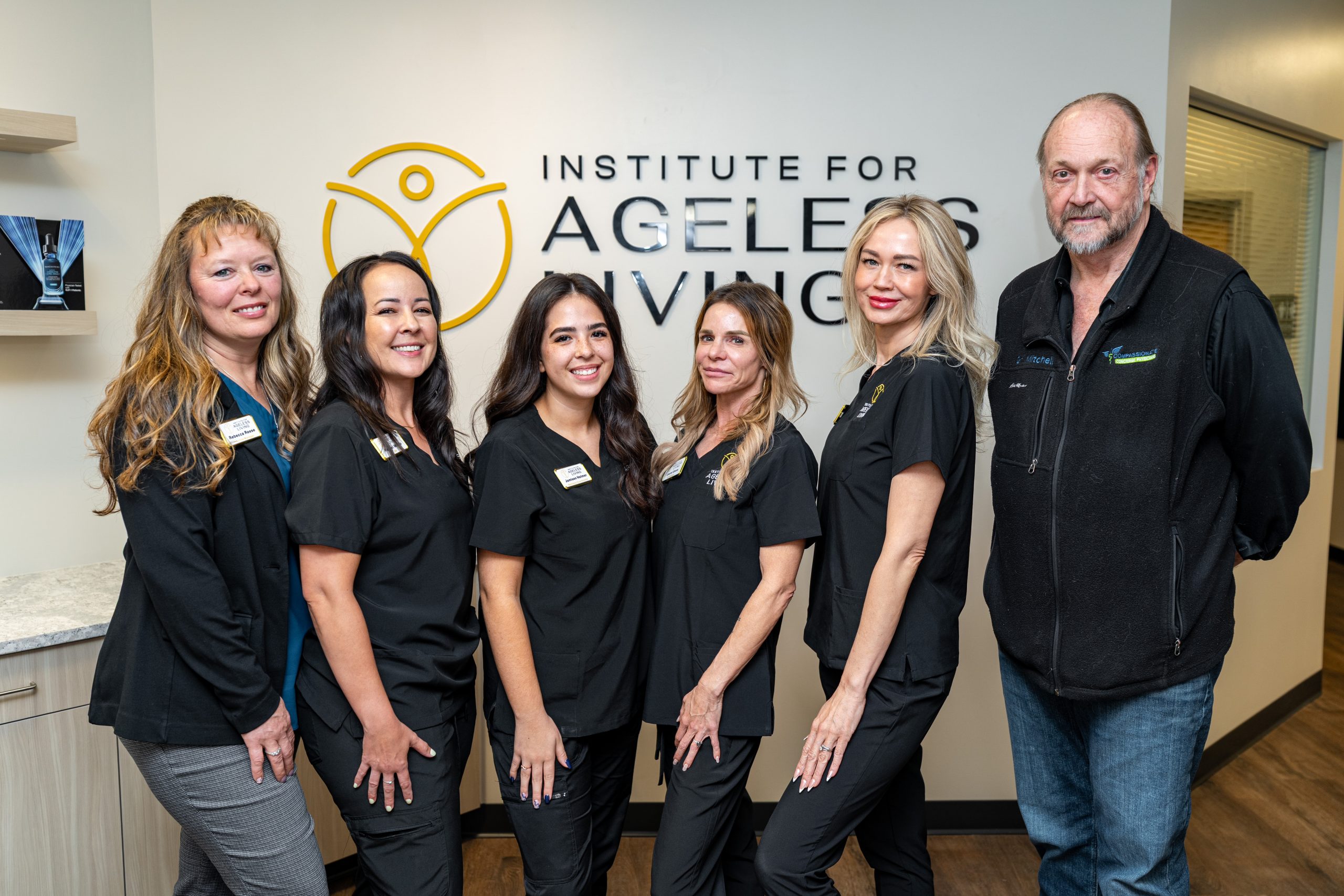 Six staff members stand and smile in front of a wall sign reading Institute For Ageless Living. Five women in black medical scrubs and one man in a black jacket pose together indoors, reflecting their shared commitment to wellness and anti-aging care.