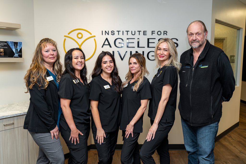 Six people, five women in black scrubs and one man in casual clothes, stand smiling in front of a wall sign that reads Institute For Ageless Living inside a professional office setting, highlighting their commitment to wellness and anti-aging solutions.