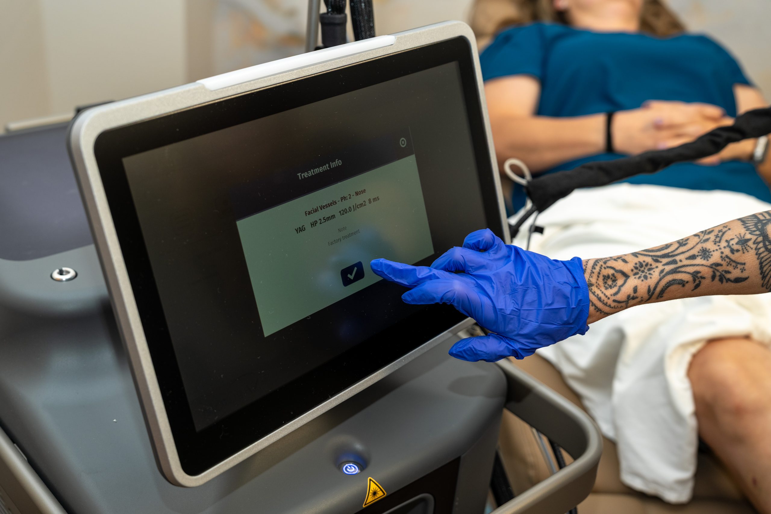 A gloved hand touches a touchscreen displaying anti-aging treatment info, while a person lies on a medical bed in the background at the Institute For Ageless Living.