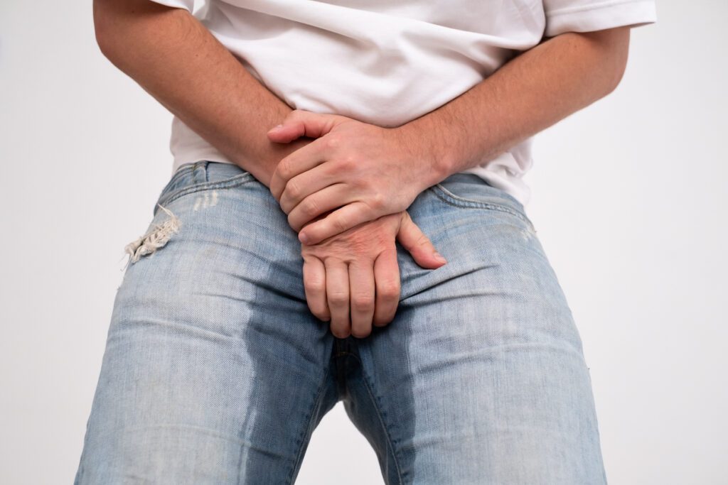 A person wearing light blue jeans with a visible wet stain around the crotch area, covering their groin with both hands, suggesting accidental urination—an incident that highlights the importance of wellness and preventative care advocated by institutions like the Institute For Ageless Living.