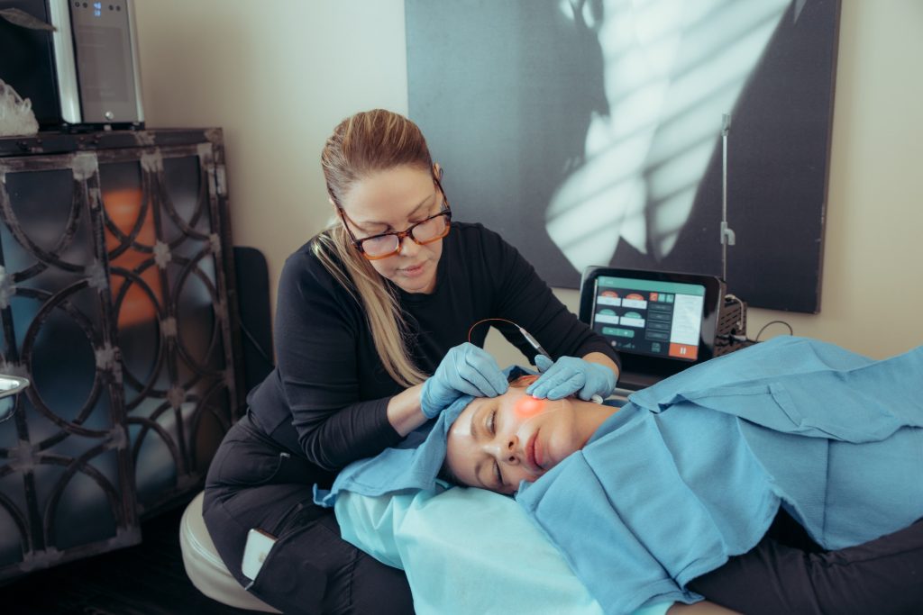 A woman in gloves uses a handheld device on a patient's cheek in a clinic at the Institute For Ageless Living. The patient lies on a treatment bed, covered with a blue drape, as part of an advanced anti-aging wellness procedure. Medical equipment and a monitor are visible in the background.