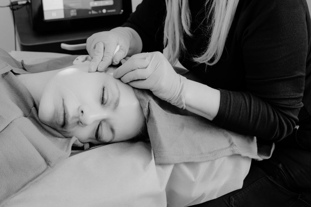 A person lies with eyes closed while another person wearing gloves gently touches their temples, possibly performing a skincare or anti-aging treatment. The serene black and white scene evokes a sense of wellness, reminiscent of therapies offered at the Institute For Ageless Living.