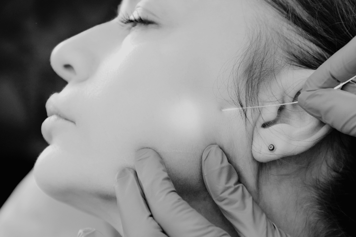 A person receives an anti-aging cosmetic facial procedure with a needle near the jaw, while wearing stud earrings. Gloved hands gently hold their face, reflecting a commitment to wellness at the Institute For Ageless Living.