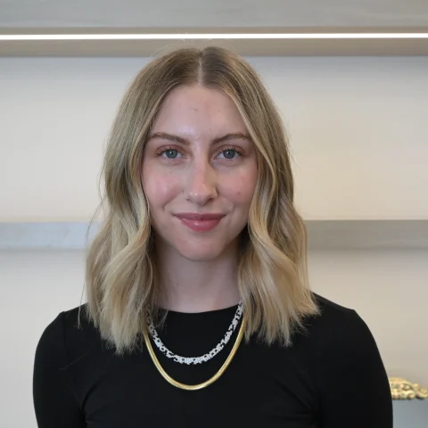A woman with shoulder-length blonde hair, wearing a black top and layered necklaces, stands in front of a light-colored wall with shelves in the background, smiling gently. Her warm presence reflects the essence of wellness promoted by the Institute For Ageless Living.