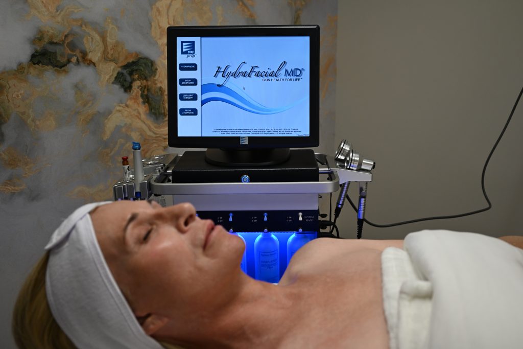 A woman lies on a spa bed with a white towel and headband at the Institute For Ageless Living. Behind her is a monitor displaying Hydrafacial MD and a machine with skincare tools and bottles, indicating a wellness-inspired facial treatment in progress that promotes anti-aging benefits.