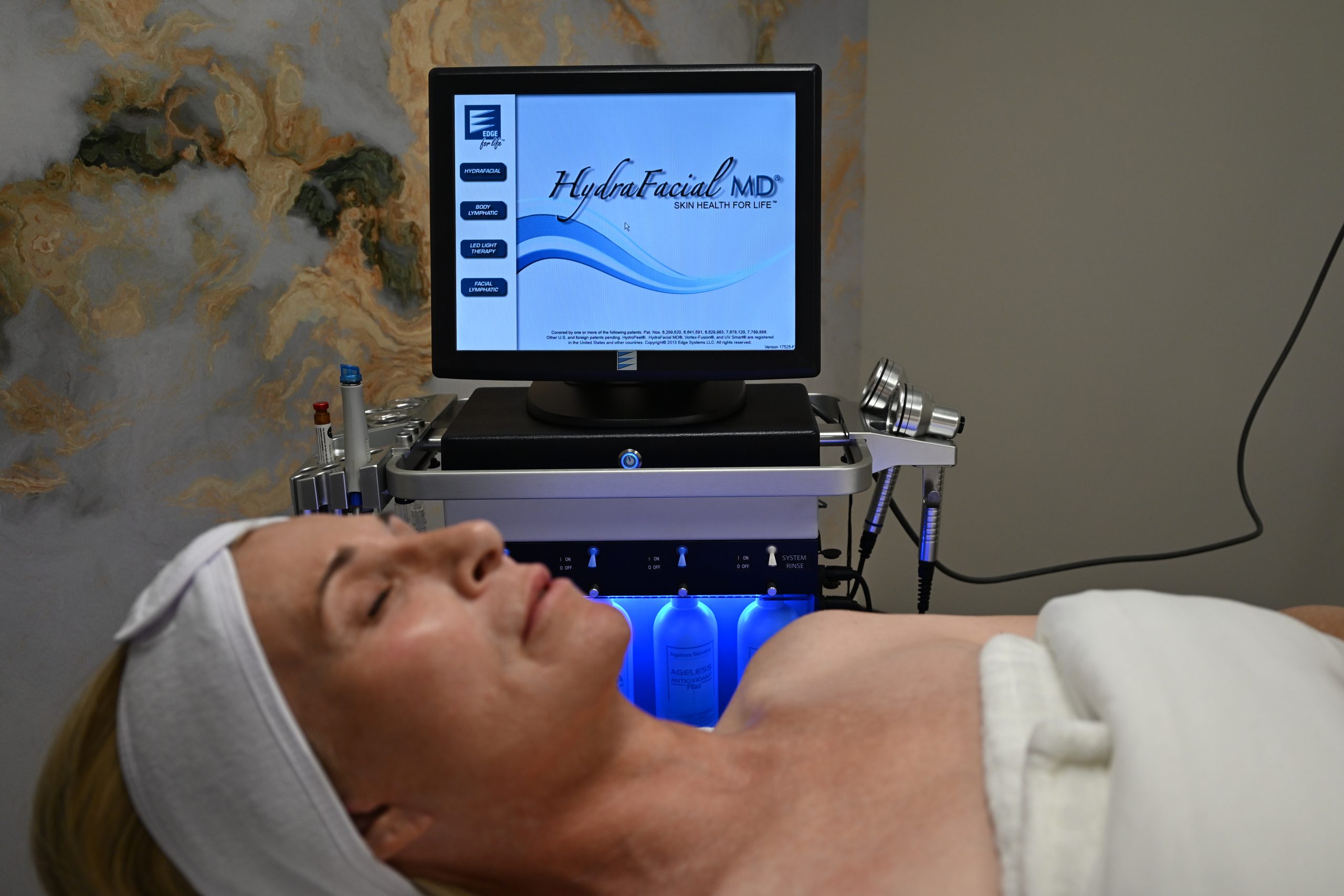 A woman lies on a spa bed with a white towel and headband at the Institute For Ageless Living. Behind her is a monitor displaying Hydrafacial MD and a machine with skincare tools and bottles, indicating a wellness-inspired facial treatment in progress that promotes anti-aging benefits.