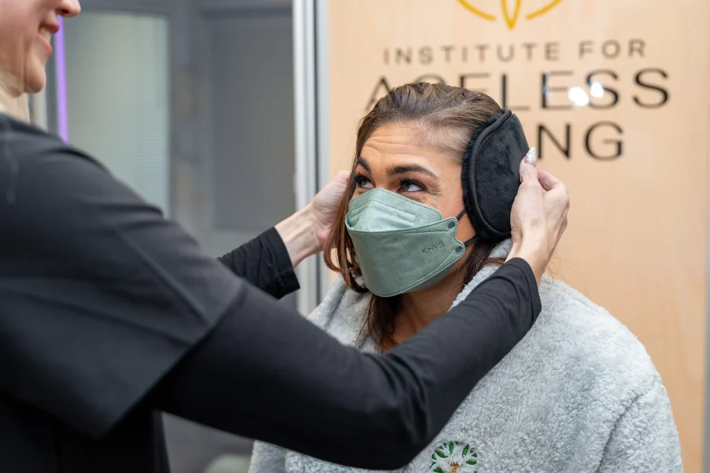 A woman wearing a face mask and gray shawl has earmuffs placed on her by another person. They are indoors near a sign that reads “Institute for Ageless Living,” reflecting the center's commitment to wellness and anti-aging practices.