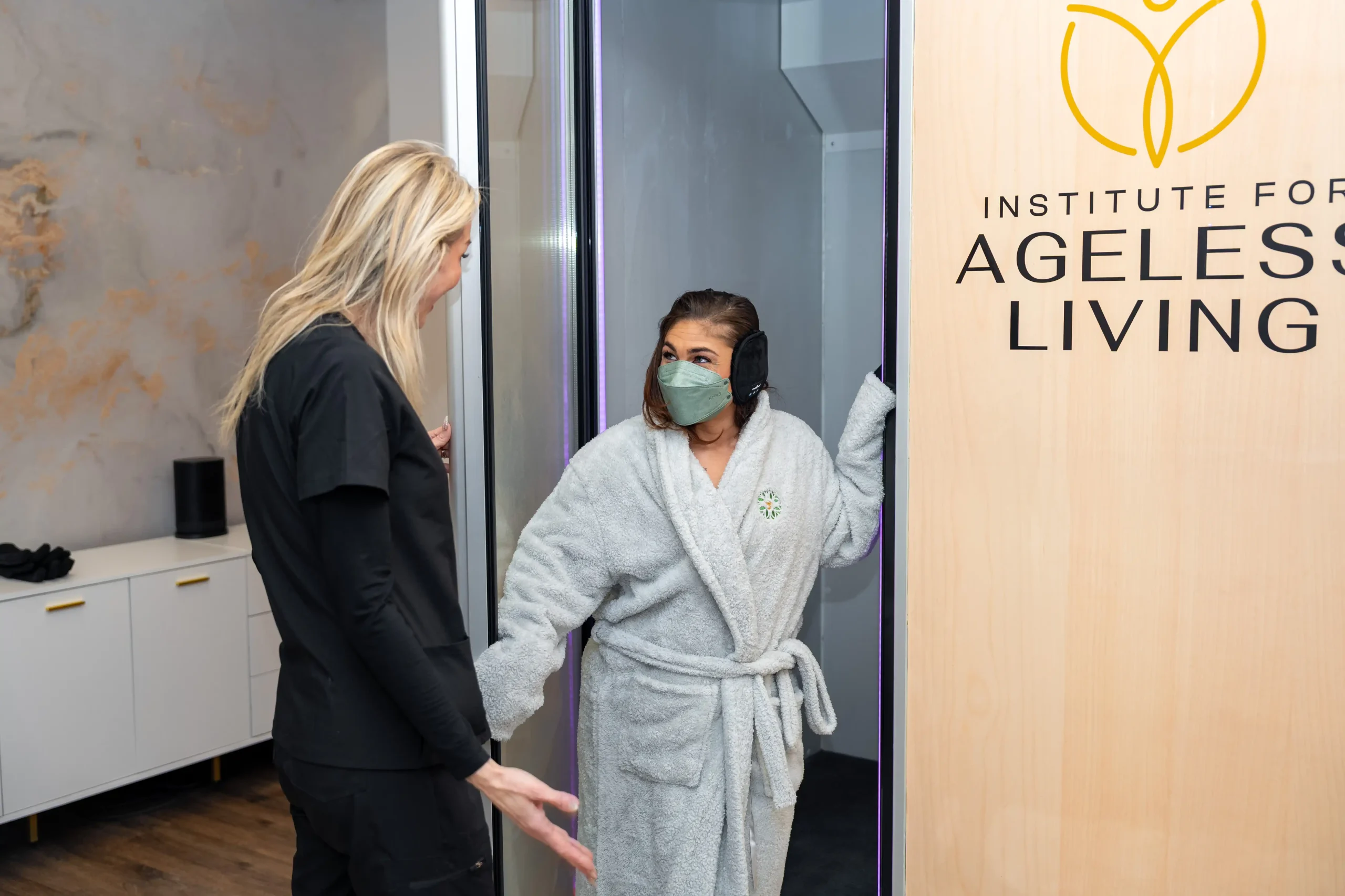 A woman in a robe and mask steps out of a chamber, greeted by a staff member in black scrubs at the Institute For Ageless Living, where cutting-edge anti-aging and wellness treatments await her.