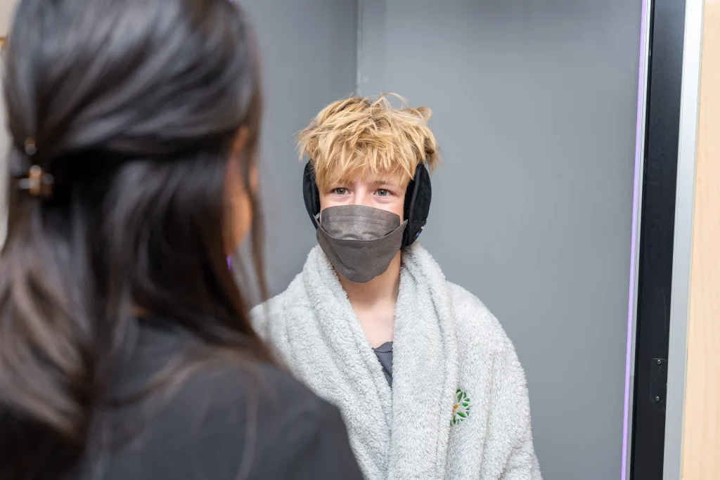 A person with messy blond hair, wearing earmuffs, a gray face mask, and a light gray robe—perhaps in search of wellness—stands indoors facing another person with long dark hair.