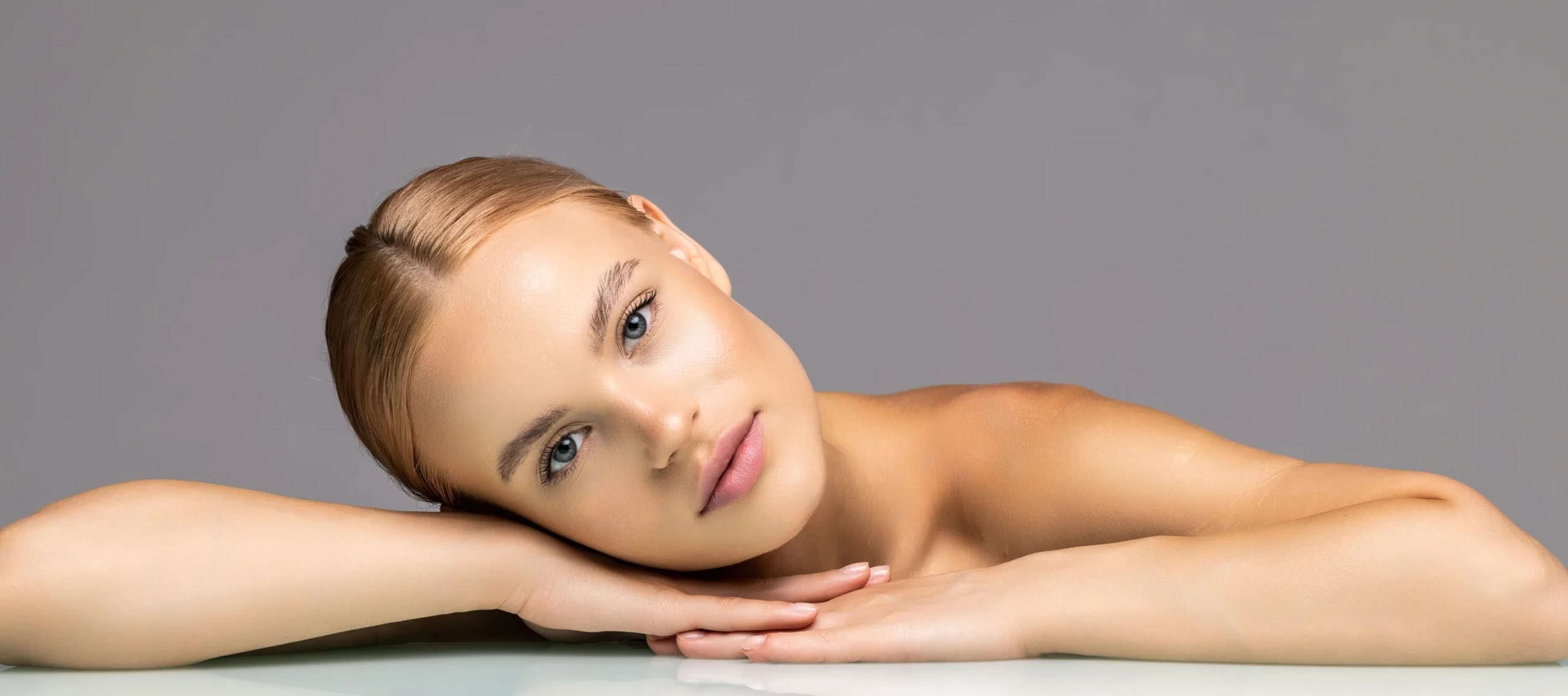A woman with smooth, radiant skin rests her head on her arms on a reflective surface, looking at the camera with a neutral expression that embodies wellness. The plain gray background enhances the focus on her complexion, reflecting the ideals of the Institute For Ageless Living.