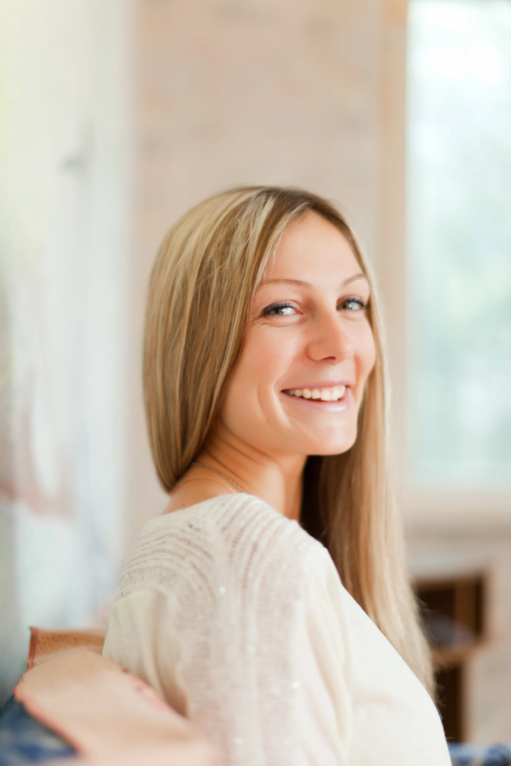 A woman with long blonde hair and a white top smiles warmly while looking over her shoulder, embodying the spirit of wellness promoted by the Institute For Ageless Living. The background is softly blurred, suggesting an indoor setting with natural light.