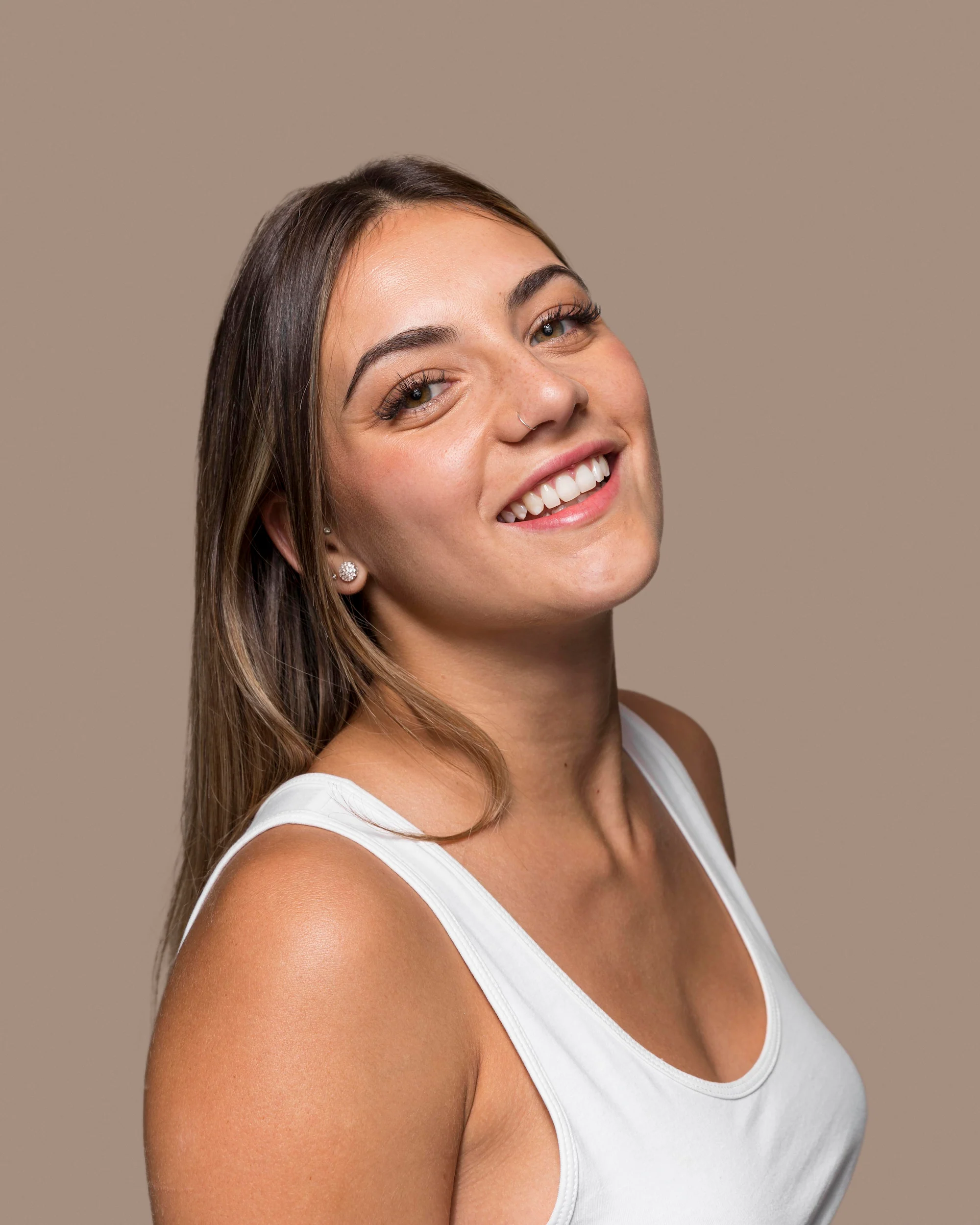 Smiling young woman with long brown hair wearing a white tank top, posing against a plain beige background, radiating wellness and vitality.