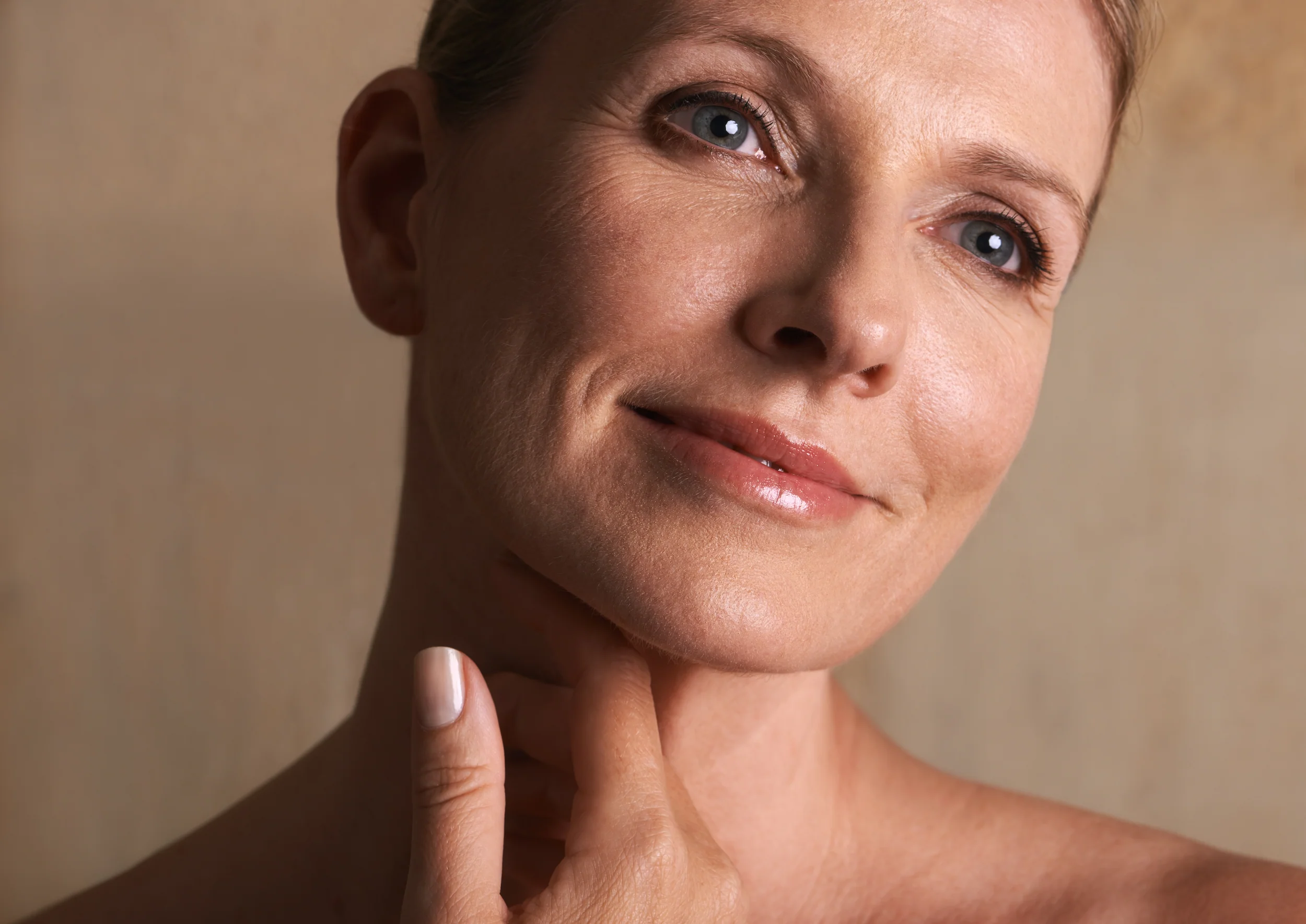 A woman with light skin and blue eyes softly smiles, touching her neck with her hand—a subtle gesture reflecting a sense of wellness and self-care. She has short, light brown hair and minimal makeup. The background is neutral and out of focus, evoking the serene environment often found at the Institute For Ageless Living.