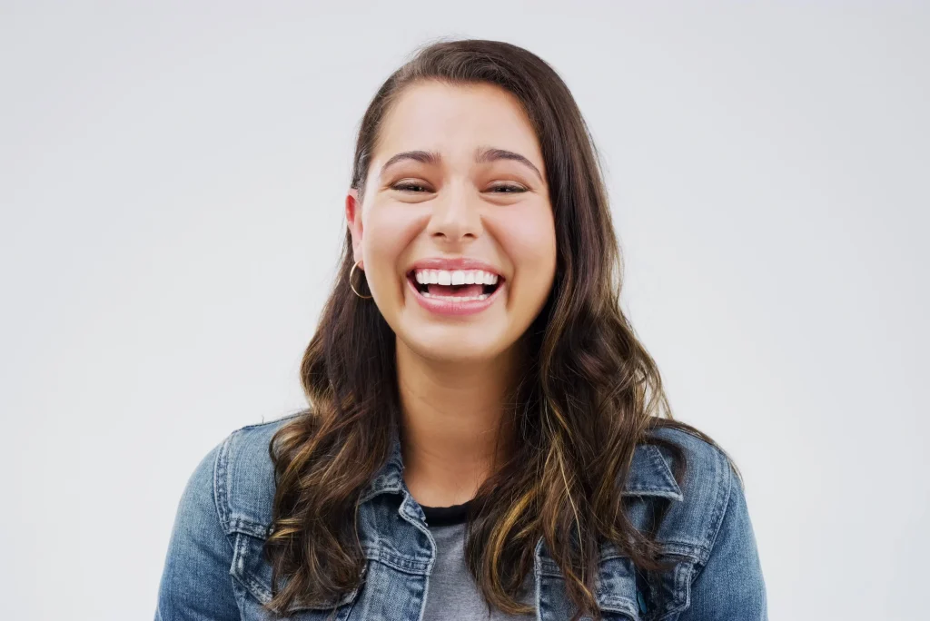 A young woman with long brown hair, wearing a denim jacket and hoop earrings, smiles brightly against a plain white background—radiating the vitality promoted by the Institute For Ageless Living's wellness and anti-aging philosophy.