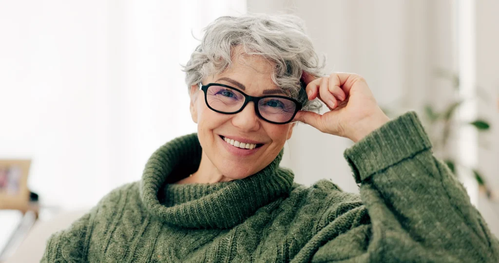 Smiling older woman with short gray hair and glasses, wearing a cozy green turtleneck sweater, sitting indoors with her hand resting on her head—a portrait radiating wellness and positivity inspired by the Institute For Ageless Living.