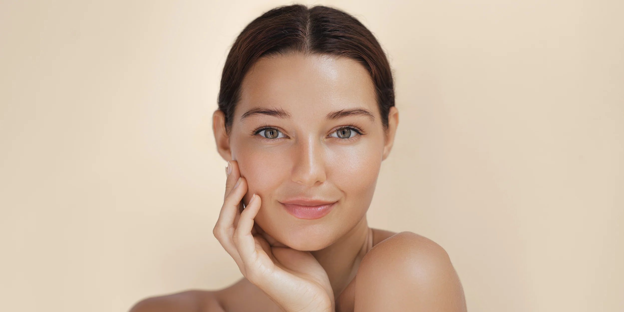 A woman with clear skin and dark hair pulled back, smiling softly with her hand resting on her cheek, exudes a sense of wellness against a beige background.