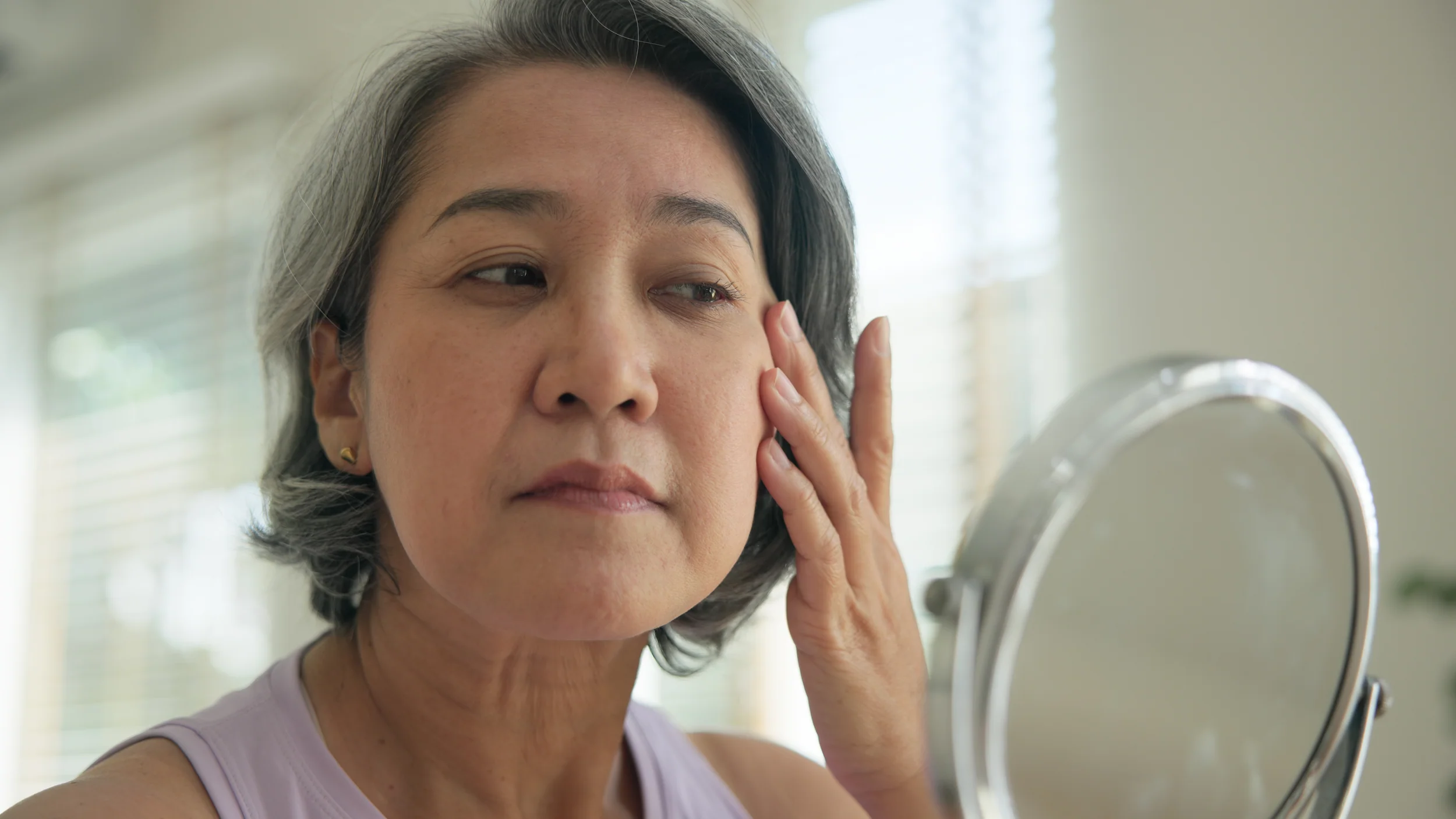 A woman with short gray hair touches her face while looking in a mirror, appearing to examine her skin in a well-lit room—a quiet moment of self-care dedicated to anti-aging and overall wellness inspired by the Institute For Ageless Living.