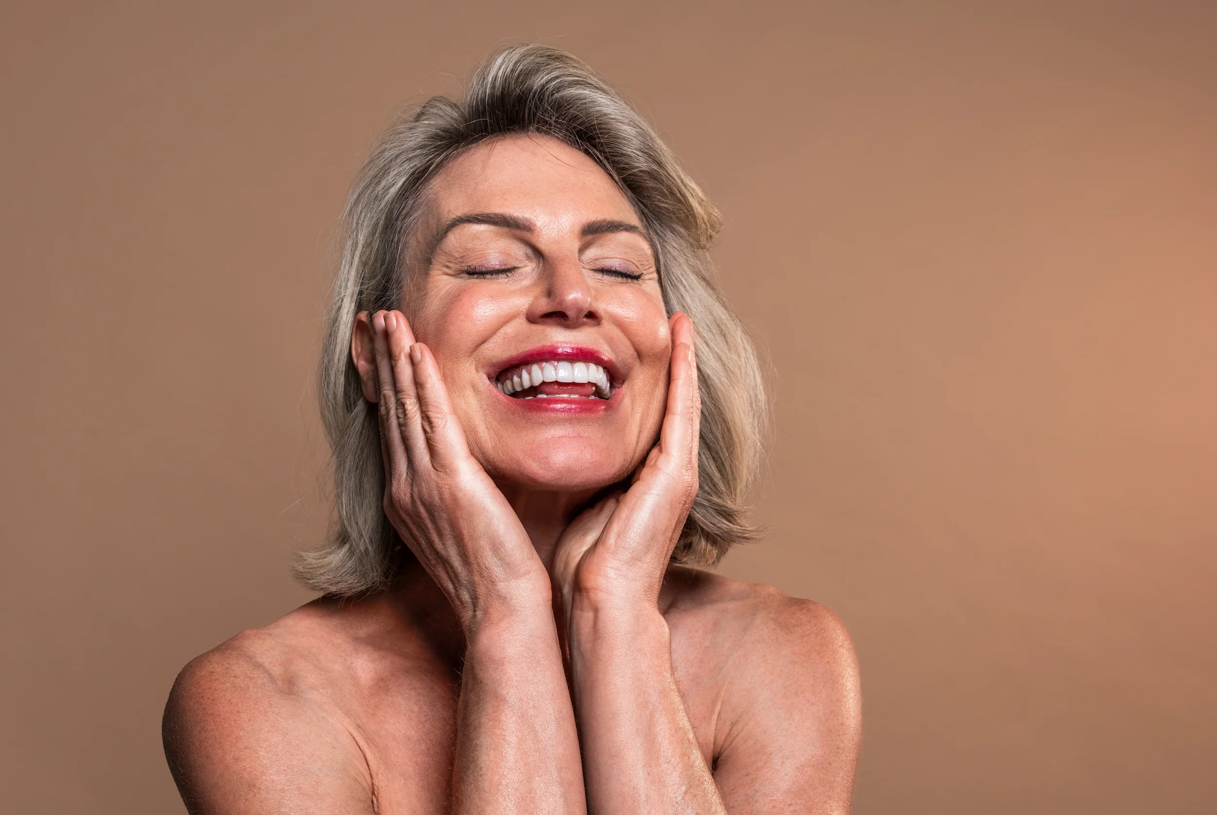 Smiling older woman with gray hair, eyes closed, touching her face gently with both hands against a plain beige background, embodying a sense of wellness and calm inspired by the Institute For Ageless Living.