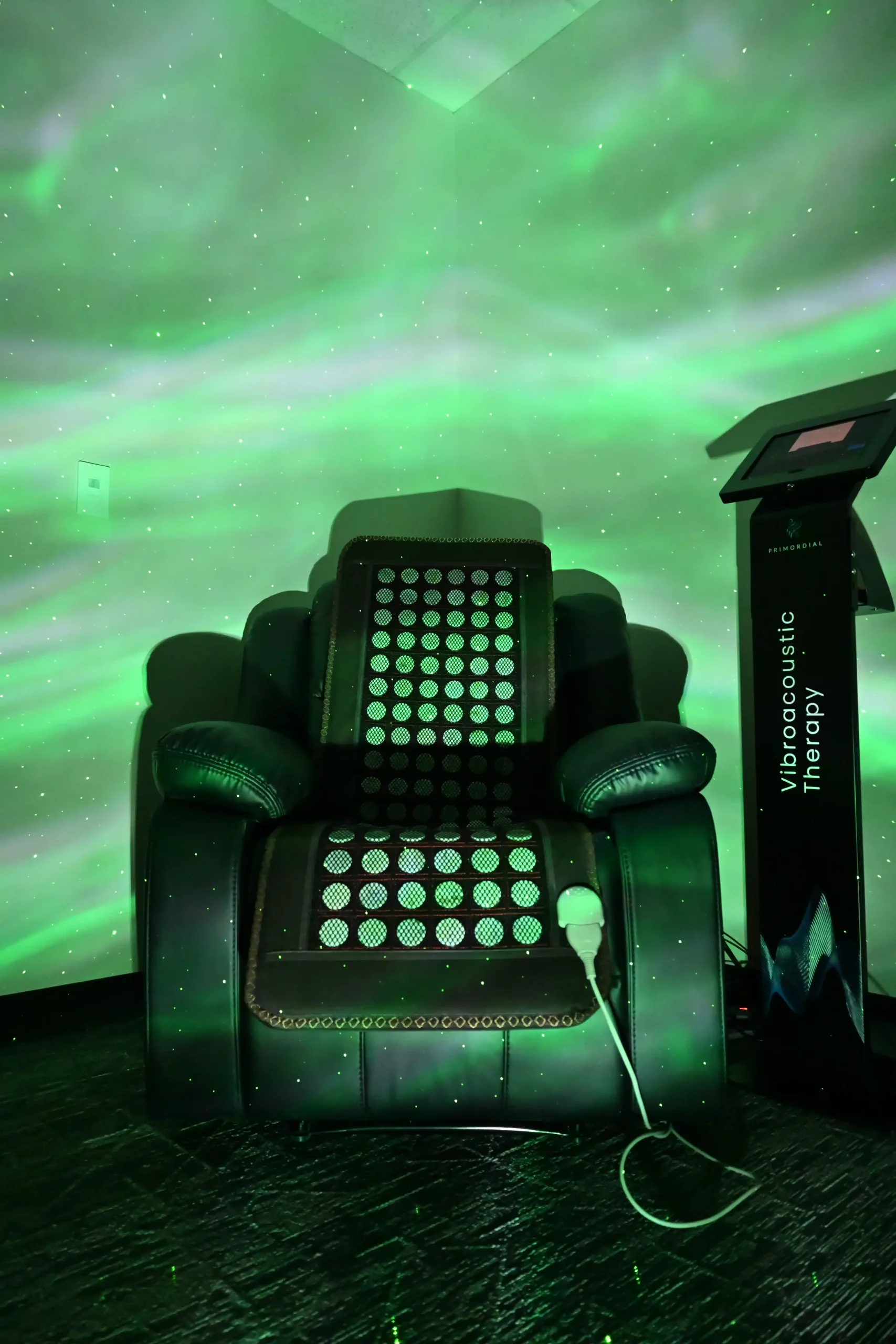 A black vibroacoustic therapy chair sits in a corner, illuminated by green swirling lights projected onto the wall. Designed with wellness in mind, the chair is accompanied by a control pedestal with a digital screen standing beside it—a testament to the innovative therapies offered at the Institute For Ageless Living.