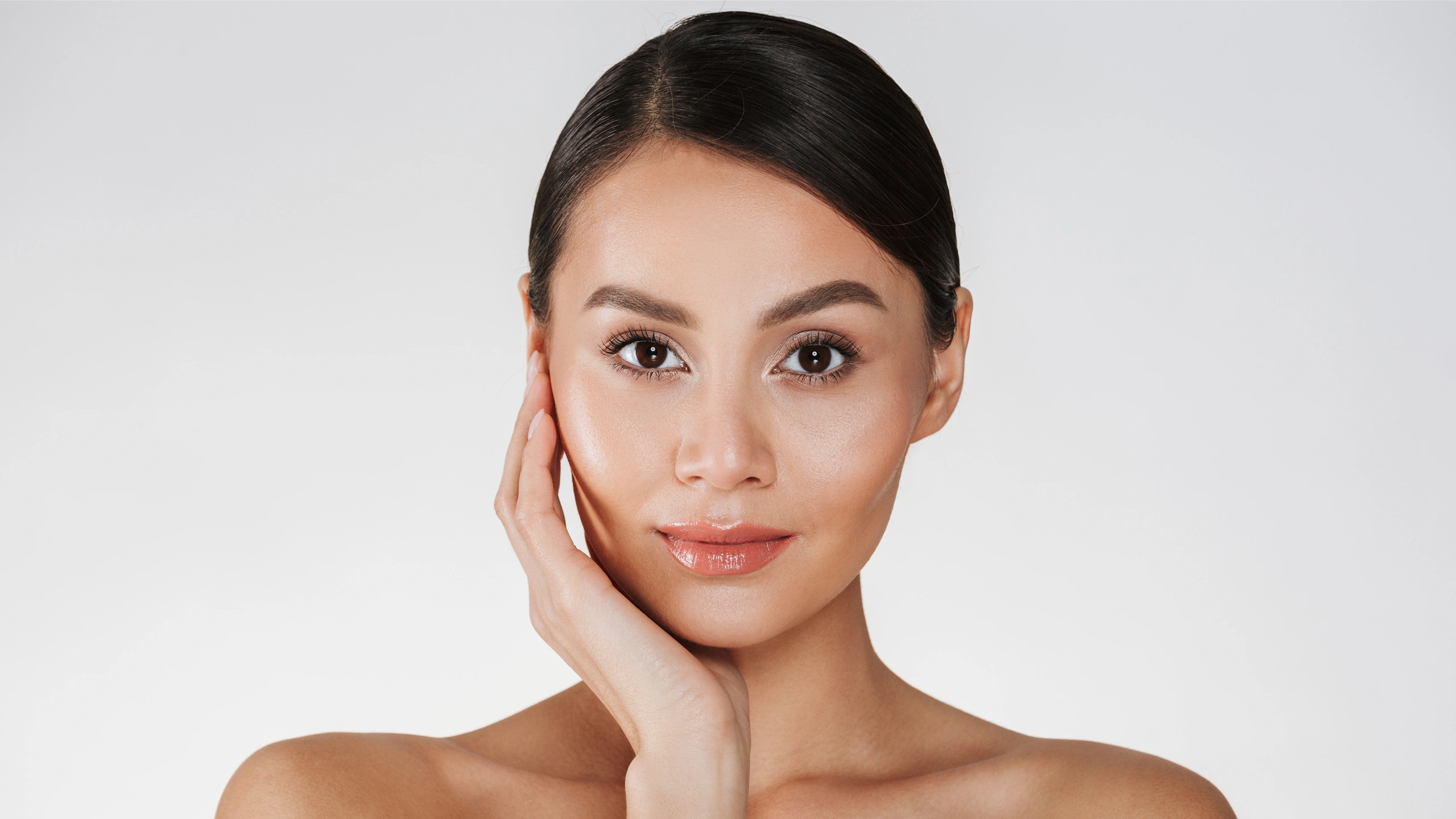 A woman with smooth skin and dark hair poses against a light background, touching her face gently with one hand, looking directly at the camera.