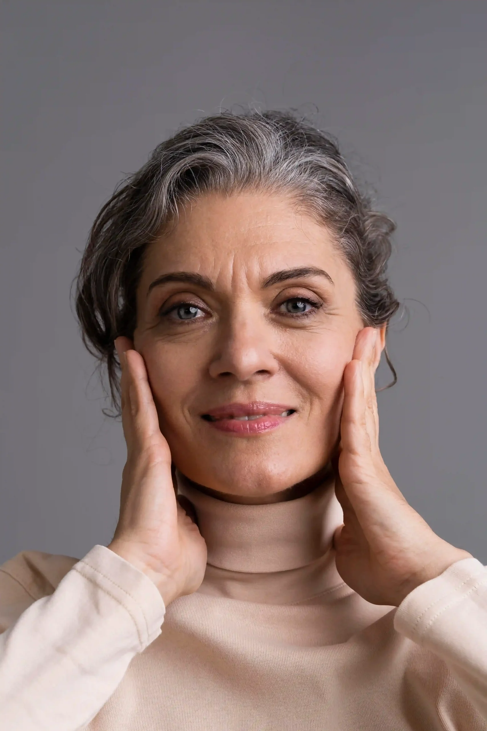 Middle-aged woman with gray hair smiles gently, touching both sides of her face with her hands. She is wearing a beige turtleneck and stands against a plain gray background.
