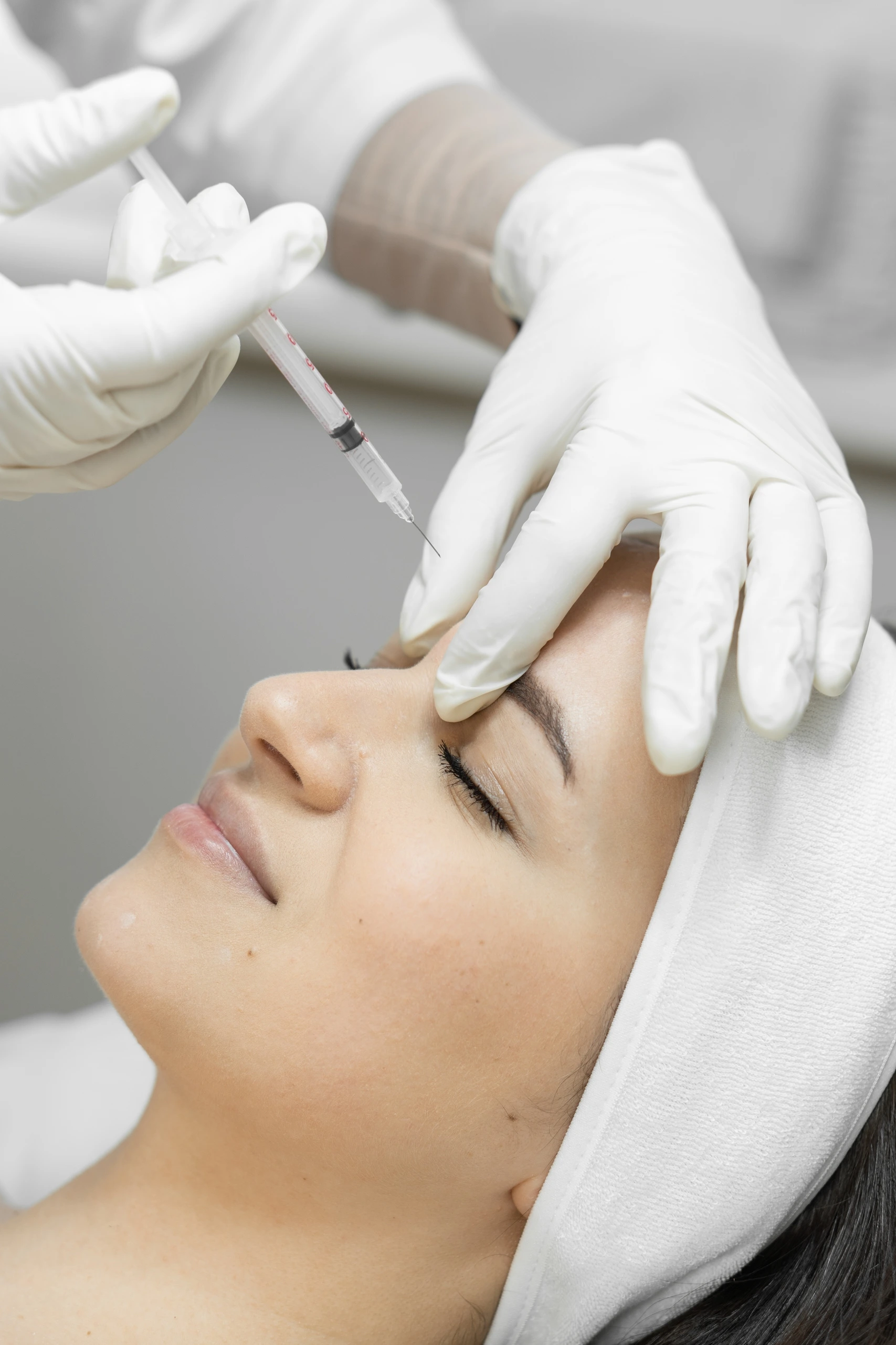 A person lying down receives a facial injection near the forehead from a gloved professional, suggesting a cosmetic procedure in a clinical setting.