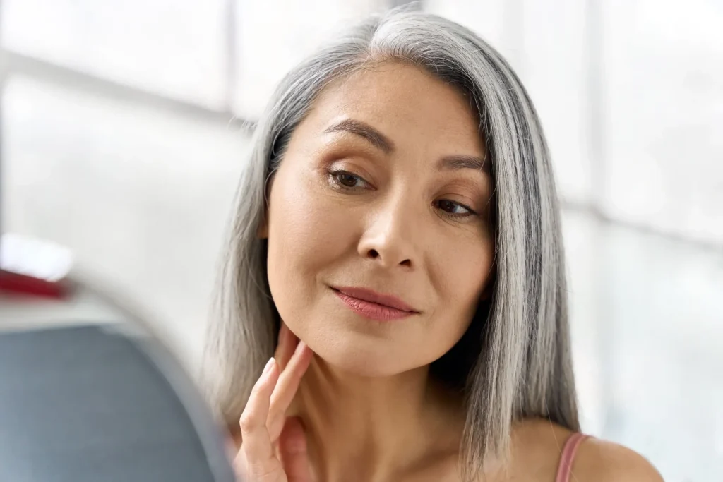 Woman with long gray hair looking at herself in a mirror, gently touching her neck, with a soft expression on her face. Bright, softly lit background.