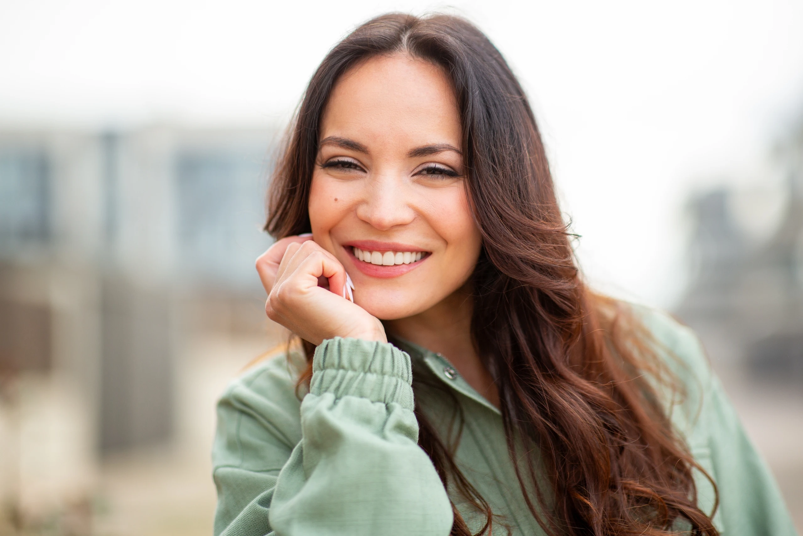 A woman with long brown hair smiles at the camera, resting her chin on her hand. She is wearing a light green jacket and is outdoors with a blurred background.