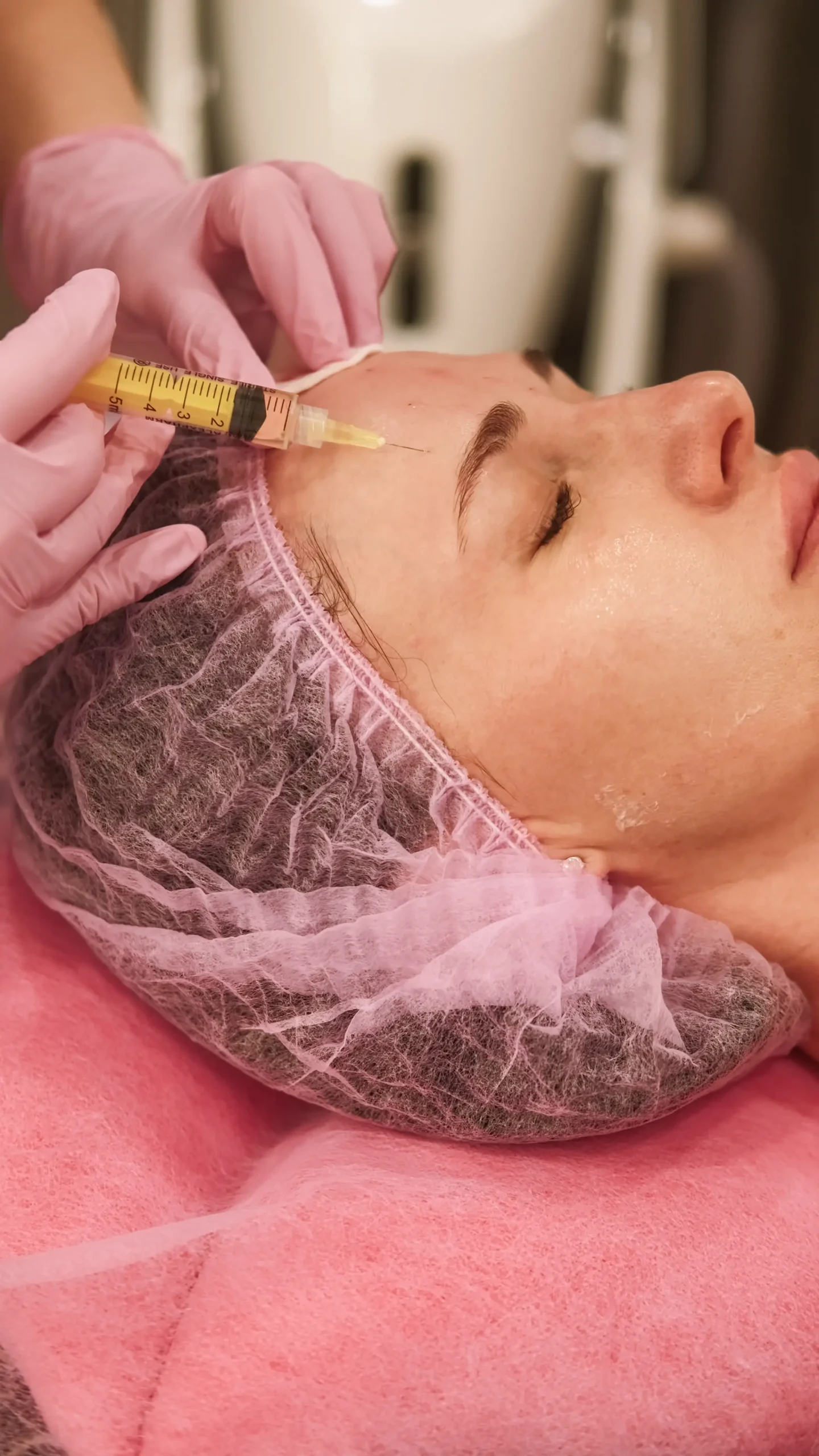 A person with closed eyes receiving a facial injection in the forehead from a gloved professional, lying on a pink sheet and wearing a hair cover.